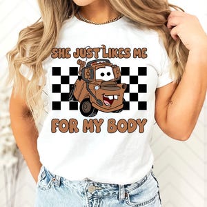 Puede incluir: Una camiseta blanca de manga corta con un gr&aacute;fico de una gr&uacute;a marr&oacute;n con auriculares, situada frente a un cuadrado de cuadros blancos y negros. El texto dice SHE JUST LIKES ME FOR MY BODY en letras marrones y redondeadas.