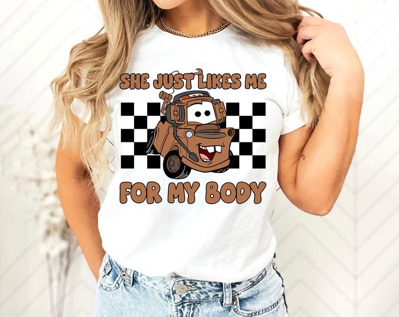 Puede incluir: Una camiseta blanca de manga corta con un gr&aacute;fico de una gr&uacute;a marr&oacute;n con auriculares, situada frente a un cuadrado de cuadros blancos y negros. El texto dice SHE JUST LIKES ME FOR MY BODY en letras marrones y redondeadas.