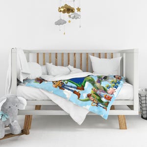 May include: A soft blanket featuring a vibrant graphic print of various Toy Story characters, including Woody, Rex, and the green aliens, set against a bright blue sky with white clouds. The blanket is draped over a white wooden crib with slats.