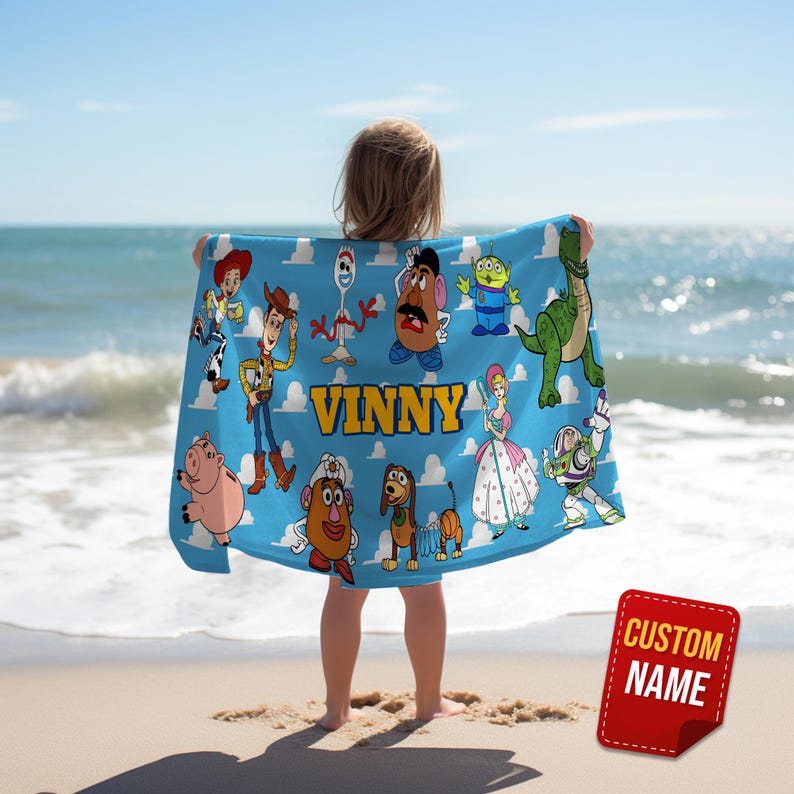 May include: A blue blanket featuring cartoon characters and the name "VINNY" in yellow letters. The blanket is held by a person on a beach. A red sticker with the words "CUSTOM NAME" is in the lower right corner.