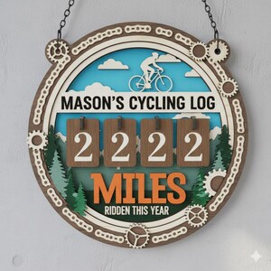 Puede incluir: Registro de ciclismo de madera con el texto "MASON'S CYCLING LOG" y "2222 MILES RIDDEN THIS YEAR." El diseño incluye una silueta de ciclista, nubes, árboles y detalles en forma de engranajes. El letrero es marrón, blanco, azul y naranja.