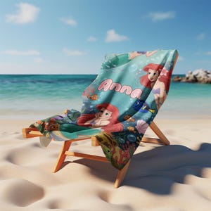 Peut inclure: Un transat avec une serviette sur le th&egrave;me de la Petite Sir&egrave;ne de couleur turquoise. La serviette repr&eacute;sente Ariel, des poissons et le nom "Anna" en &eacute;criture rose. La chaise est en bois clair et est pos&eacute;e sur une plage de sable avec l'oc&eacute;an en arri&egrave;re-plan.