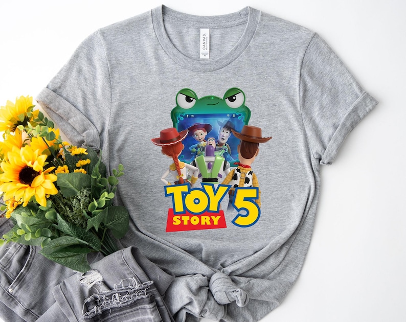 May include: A heather gray t-shirt featuring a colorful graphic from the movie Toy Story 5. The graphic includes characters like Woody, Buzz Lightyear, and Jessie, with the text "TOY STORY 5" in yellow and red.
