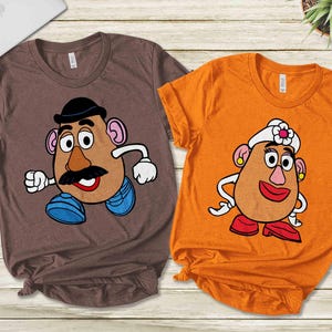 May include: Two t-shirts featuring cartoon potato head characters. One is brown with a male character wearing a hat, mustache, and blue shoes. The other is orange with a female character wearing a hat and red shoes.