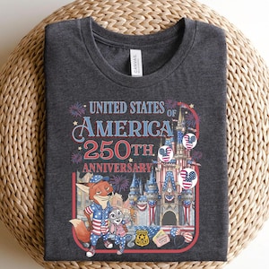 Puede incluir: Camiseta gris oscuro con un gr&aacute;fico colorido que celebra el 250 aniversario de los Estados Unidos de Am&eacute;rica. El dise&ntilde;o incluye fuegos artificiales, un castillo y personajes de dibujos animados. El texto "UNITED STATES OF AMERICA 250TH ANNIVERSARY" es visible.