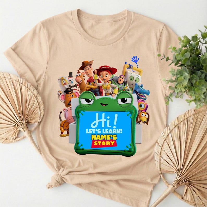 May include: A beige t-shirt with a vibrant graphic of animated characters and a green frog-shaped sign. The sign displays "Hi! LET'S LEARN! NAME'S STORY" in white and yellow text. The shirt is made of a soft material.