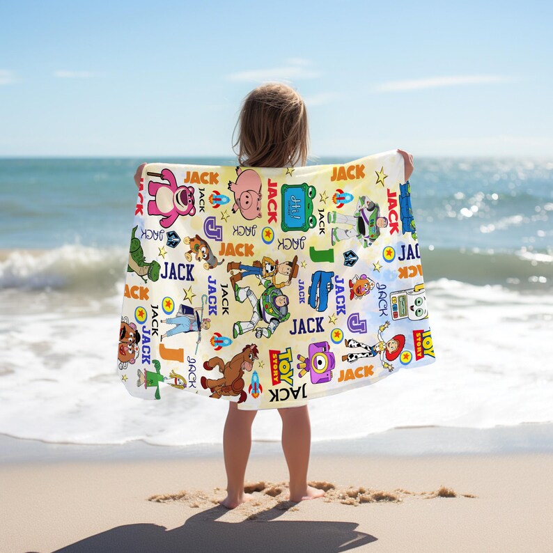 May include: A beach towel featuring various Toy Story characters and the name "JACK" repeated throughout. The towel has a light yellow background with colorful illustrations of Woody, Buzz Lightyear, and other characters. The towel is held up by a person on a sandy beach.