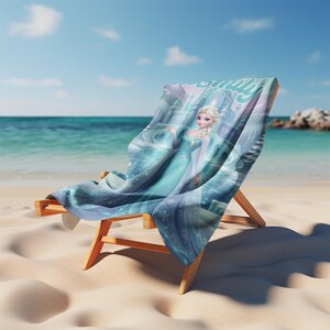 May include: A wooden beach chair draped with a rectangular towel featuring a graphic print of a blonde character in a light blue gown, standing before a castle. The towel displays swirling blue patterns and the name Emily in cursive script.