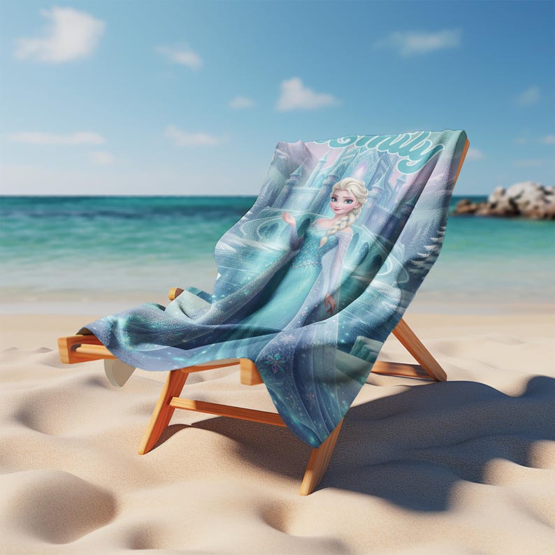 May include: A wooden beach chair draped with a rectangular towel featuring a graphic print of a blonde character in a light blue gown, standing before a castle. The towel displays swirling blue patterns and the name Emily in cursive script.
