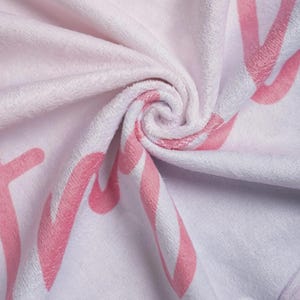 Puede incluir: Una manta de tela de felpa blanca suave con un patr&oacute;n repetitivo de letras grandes en cursiva de color rosa. El material tiene una textura difusa similar al terciopelo con pliegues profundos que crean un efecto giratorio tridimensional.
