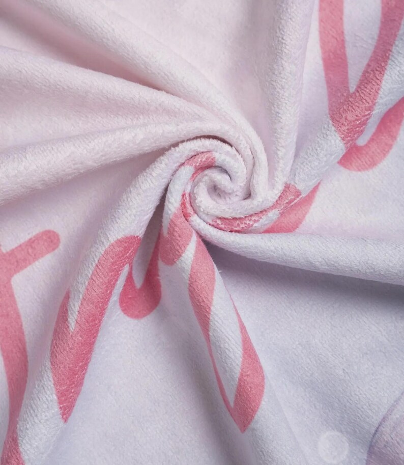 Puede incluir: Una manta de tela de felpa blanca suave con un patr&oacute;n repetitivo de letras grandes en cursiva de color rosa. El material tiene una textura difusa similar al terciopelo con pliegues profundos que crean un efecto giratorio tridimensional.