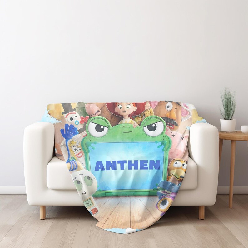 May include: A soft fleece blanket featuring a colorful graphic print of various animated characters surrounding a central green frog-shaped frame. The word ANTHEN is printed in bold blue capital letters inside the center of the green frog frame.