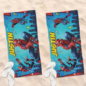 Op de afbeelding: Twee blauwe strandlakens met Spider-Man-illustraties in actieposes. De lakens hebben een lichtblauwe achtergrond met stadsgezichten, bliksemflitsen en de naam "JUSTIN" in geel. Witte teenslippers zijn bij de lakens geplaatst.