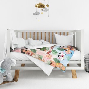 Puede incluir: Una manta para beb&eacute; con una colorida ilustraci&oacute;n de una mujer polinesia, un cerdo y una vaca. La manta tiene un fondo rosa y azul con hojas tropicales y la palabra "Nam" en blanco.