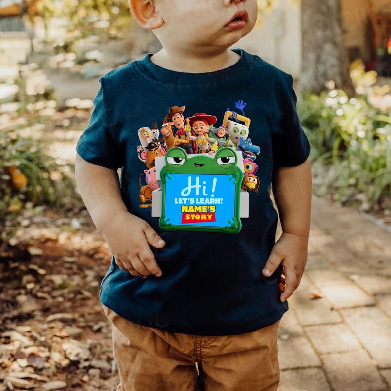 May include: Navy blue toddler t-shirt with a colourful graphic of animated film characters. The graphic includes the text "Hi! Let's Learn! Name's Story" on a frog-shaped sign. The shirt is worn with brown trousers.