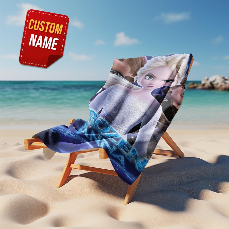 May include: A beach chair with a blue and white patterned towel featuring a cartoon character. The chair is made of wood and is on a sandy beach with the ocean in the background. A red tag with the text "CUSTOM NAME" is in the upper left corner.
