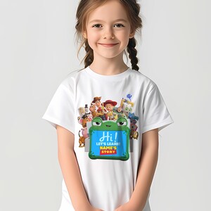 May include: White t-shirt with a colourful graphic of animated characters and a green frog-shaped sign. The sign reads "Hi!" and "Let's Learn! NAME'S STORY" in blue and yellow text. The shirt is designed for children.