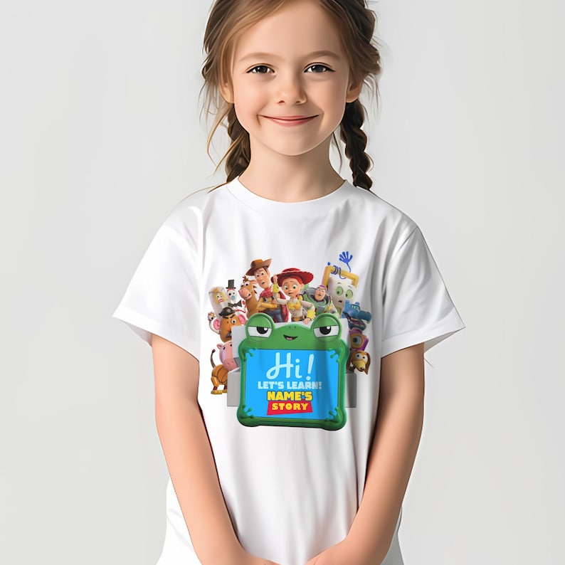 May include: White t-shirt with a colourful graphic of animated characters and a green frog-shaped sign. The sign reads "Hi!" and "Let's Learn! NAME'S STORY" in blue and yellow text. The shirt is designed for children.