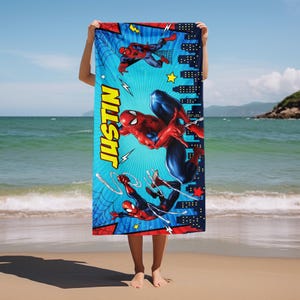Op de afbeelding: Een rechthoekige strandlaken met Spider-Man in actieposes. De handdoek heeft een blauwe achtergrond met de naam "JUSTIN" in geel. Spider-Man is afgebeeld in zijn klassieke rood-blauwe pak, met stads- en webontwerpen.