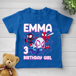 May include: A royal blue short-sleeve t-shirt featuring a graphic print of three cartoon spider characters against a pink spiderweb background. The text reads EMMA, 3, and BIRTHDAY GIRL in white block letters with a pink outline on the front.