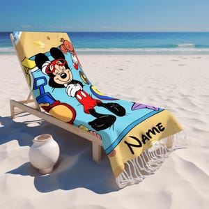 May include: A beach towel featuring a cartoon Mickey Mouse design, lying on a wooden beach chair. The towel has a light blue background with Mickey Mouse in red shorts and sunglasses. The towel has a yellow border with the word "Name" and white fringe.