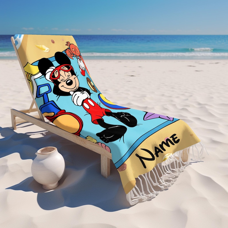 May include: A beach towel featuring a cartoon Mickey Mouse design, lying on a wooden beach chair. The towel has a light blue background with Mickey Mouse in red shorts and sunglasses. The towel has a yellow border with the word "Name" and white fringe.