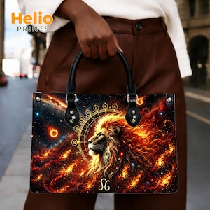 May include: A rectangular handbag featuring a vibrant cosmic print of a lion's head surrounded by fiery nebula clouds, stars, and a golden zodiac circle. The bag has two black handles attached with silver-toned hardware and a gold Leo symbol at the base.