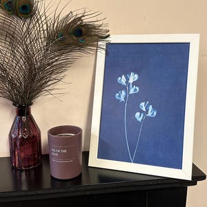 May include: A white framed print of a blue flower on a dark blue background. The print is sitting on a black shelf with a brown candle that says "All of the Love" on it. A peacock feather is in the background.