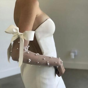 May include: A strapless white wedding dress with a fitted bodice and a flowing skirt. The dress features sheer, long sleeves adorned with scattered pearls. A large white bow accents the upper arm, adding a touch of elegance to the bridal gown.