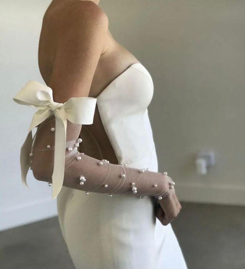 May include: A strapless white wedding dress with a fitted bodice and a flowing skirt. The dress features sheer, long sleeves adorned with scattered pearls. A large white bow accents the upper arm, adding a touch of elegance to the bridal gown.