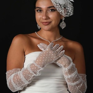 May include: A woman wearing a white strapless dress, a dotted veil, and long white gloves with a dotted pattern. She is also wearing a diamond necklace and earrings. The background is black.