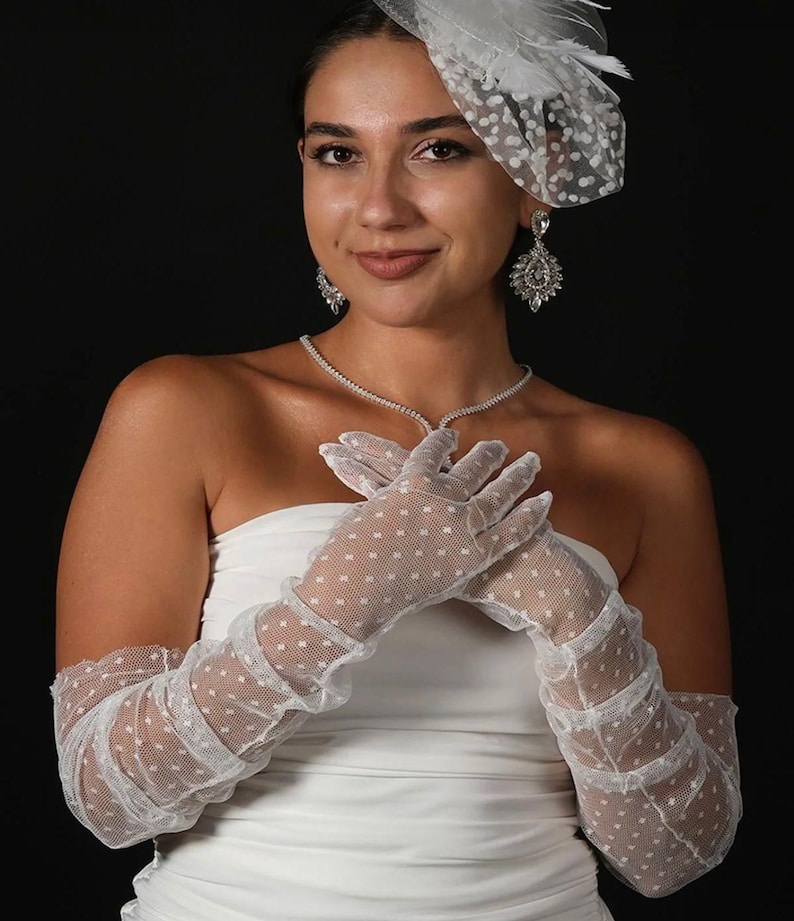 May include: A woman wearing a white strapless dress, a dotted veil, and long white gloves with a dotted pattern. She is also wearing a diamond necklace and earrings. The background is black.