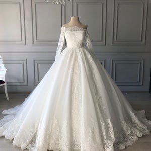 Peut inclure: Une robe de mariée blanche avec un col bateau et des manches longues. La robe présente des détails de dentelle complexes sur le corsage, les manches et l'ourlet, avec une jupe ample et une traîne.