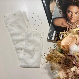 May include: White sheer gloves adorned with scattered faux pearls. The gloves are laid out on a white surface, with a fashion magazine and a floral arrangement in the background. The gloves appear to be for a special occasion.