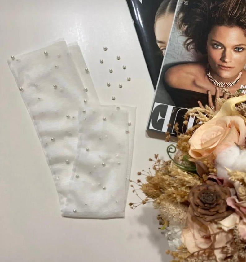 May include: White sheer gloves adorned with scattered faux pearls. The gloves are laid out on a white surface, with a fashion magazine and a floral arrangement in the background. The gloves appear to be for a special occasion.