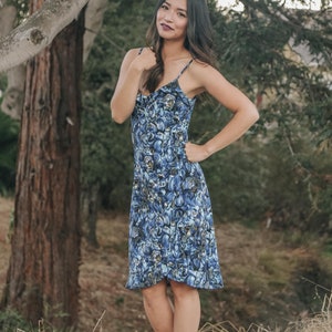 May include: A woman wearing a blue and white floral print midi dress with a spaghetti strap neckline. She is standing in a grassy area with trees in the background.