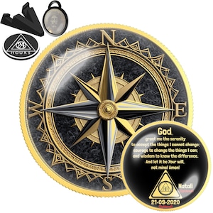 May include: A round gold-rimmed medallion featuring a black compass rose design with cardinal directions N, S, E, W. A smaller circular insert displays the Serenity Prayer text, a triangle logo with 24 HOURS, a date 21-09-2020, and a small oval Jesus pendant.