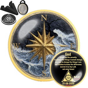 May include: A round gold-rimmed medallion featuring a compass rose over a dark ocean wave design. A smaller circular insert displays the Serenity Prayer text, a triangle symbol with 24 hours, and a date. Includes a black stand and a small pendant.