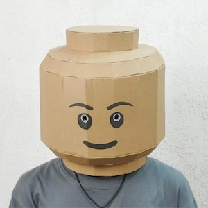 Könnte beinhalten: Eine Person trägt eine große, handgefertigte Lego-Kopfmaske aus Pappe. Die Maske ist beige mit schwarz bemalten Augen und einem Lächeln. Die Person trägt ein graues T-Shirt und eine Halskette.