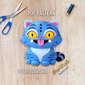 Puede incluir: Un gato de fieltro azul con ojos amarillos, lengua rosa y dientes blancos. El gato está rodeado de materiales de costura, incluyendo tijeras e hilo. El texto en la imagen dice "PDF PATTERN" y "3 SIZE AVAILABLE".