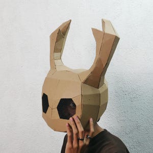 Puede incluir: Una máscara de cartón marrón con un diseño geométrico, con cuernos grandes y estilizados y aberturas para los ojos con malla negra. La máscara está hecha a mano y parece ser usada por una persona.
