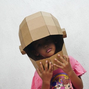 Puede incluir: Un niño con un gran casco de cartón marrón. El casco está hecho de piezas de cartón dobladas, creando una forma geométrica. El niño lleva una camiseta rosa con un diseño de unicornio.