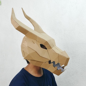 Peut inclure: Un masque de dragon en carton brun avec de grandes cornes et un design géométrique détaillé. Le masque couvre toute la tête et présente des ouvertures pour les yeux et une mâchoire détaillée. Le masque est porté par une personne portant un t-shirt bleu.