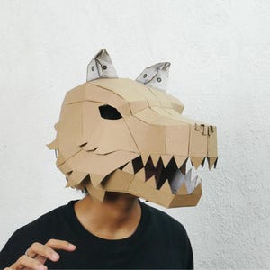 Puede incluir: Una máscara geométrica de cartón con forma de cabeza de dragón. La máscara es principalmente de color marrón claro con detalles blancos en las orejas. La boca del dragón está abierta, revelando afilados dientes. La persona lleva una camiseta negra.
