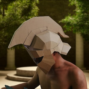 Könnte beinhalten: Ein hellbrauner Spartanischer Helm aus Pappe mit geometrischem Design. Der Helm hat einen Kamm und einen Gesichtsschutz, der aus gefalteten und geschnittenen Pappstücken besteht. Der Hintergrund ist eine verschwommene Außenszene mit Bäumen und Säulen.