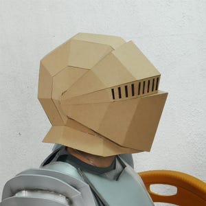 Puede incluir: Un casco de caballero de cartón beige con un diseño geométrico. El casco tiene una visera con ranuras verticales. La persona lleva una pieza de pecho plateada. Hay un objeto naranja en el fondo.