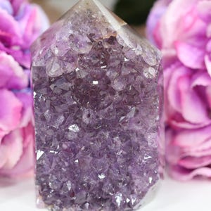 Könnte beinhalten: Eine polierte Amethyst-Kristallspitze mit einer tiefvioletten, facettierten Oberfläche und einer spitzen Oberseite. Der Kristall zeigt eine dichte Ansammlung kleiner, gezackter, durchscheinender violetter Quarzformationen, die das Licht reflektieren.