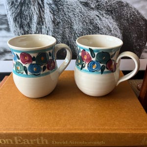 May include: Two ceramic mugs with a cream base and a floral design in blue, red, and green. Each mug has a handle and a turquoise stripe near the top. The mugs are sitting on a brown book.