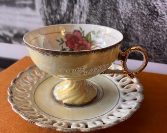 May include: A vintage teacup and saucer set with a delicate floral design in shades of pink, yellow, and green. The cup has a gold rim and a scalloped saucer.