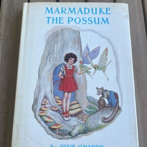May include: Vintage children's book titled "Marmaduke the Possum" by Pixie O'Harris. The cover features a colorful illustration of a girl in a red dress, fairies, and a possum. The book is set against a backdrop of a tree and mushrooms.